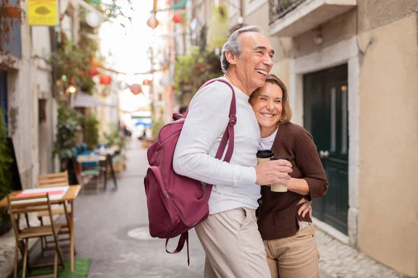 Happy European Senior Couple Traveling Standing And Hugging On Lisbon Street Outside, Holding Paper Coffee Cups. Mature Tourists Enjoying Vacation In Europe. Retirement Travel Offer
