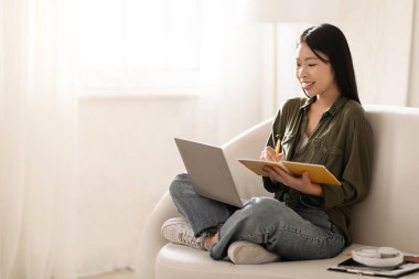 Mutlu neşeli, güzel Asyalı bir kadın, günlük öğrenci koltuğunda oturuyor, Webinar 'a katılıyor, dizüstü bilgisayar ekranına bakıyor ve notlar alıyor, fotokopi çekiyor. Çevrimiçi eğitim, e-öğrenim