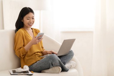 Gadget bağımlılığı. Memleketten çalışan neşeli, genç, güzel Japon kadın serbest çalışan, kanepede oturan, dizüstü bilgisayar kullanan ve akıllı telefondan mesajları kontrol eden,