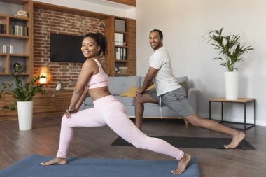 Spor giyimli genç siyah atletler birlikte bacak egzersizi yapıyorlar, oturma odasının içinde aktif ve zinde olmanın keyfini çıkarıyorlar. Esneme, vücut bakımı, zayıflama ve evdeki kaslar için uygunluk.