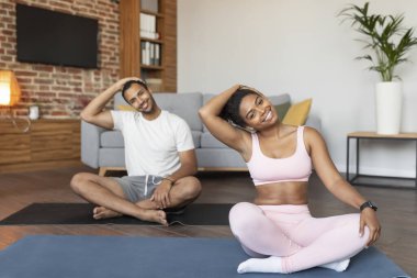 Gülümseyen siyahi erkek ve bayan spor kıyafetleri içinde egzersiz yapıyor, yoga yapıyor, oturma odasının içinde lotus pozisyonunda kaslarını esnetiyorlar. Spor, sağlık ve sağlık hizmetleri.