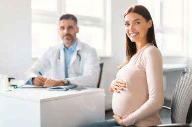 Orta yaşlı erkek jinekologla görüşürken klinikte oturan mutlu, genç hamile bir kadın göbeğini kucaklıyor ve kameraya gülümsüyor.