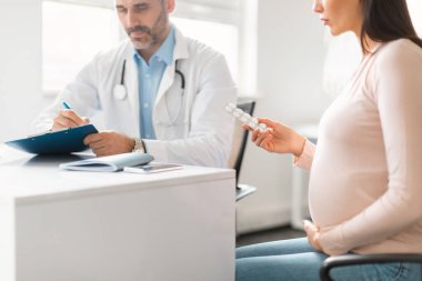 Hamilelik tedavisi. Genç hamile kadın için ilaç yazan erkek doktor tabletli su toplayan kadın hasta, klinikte oturuyor, seçici odaklanma.