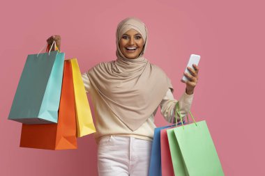 Çevrimiçi Alışveriş Konsepti. Neşeli Müslüman Kadın Akıllı Telefon ve Parlak Kağıt Paketleri, İslami Kadın Satış ve İnternet Alışverişlerinden Hoşlanıyor, Pembe Arkaplanda Duruyor, Boşluğu Kopyala