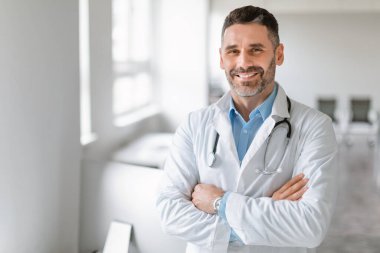 Steteskoplu iş elbisesi giyen, kliniğin içinde kollarını kavuşturmuş kameraya bakıp gülümseyen, beleş uzay boşluğu olan orta yaşlı Avrupalı doktor portresi.