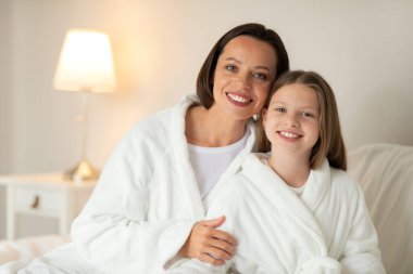 Bornozlu küçük beyaz kızı kucaklayan mutlu genç bir kadının portresi, beyaz yumuşak bir yatakta oturur, yatak odasının içinde boş zamanların ve güzellik bakımının keyfini çıkarır. Aile sevgisi, anne kız ilişkisi.