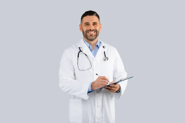 白いコートを着た幸せな中年の男性医師は 聴診器を持ち クリップボードを保持し カメラで笑顔 灰色の背景にポーズをとります 医療サービス セラピストとの検査 — ストック写真