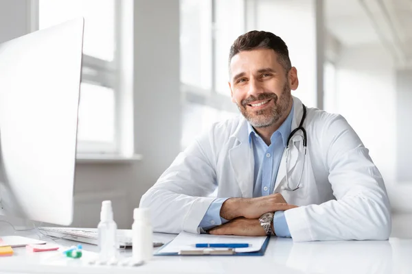 穿着白色医疗制服的快乐男医生的画像 坐在医院的办公桌前 对着相机微笑 自由自在的空间 男科医生或在私人诊所工作的医生 — 图库照片