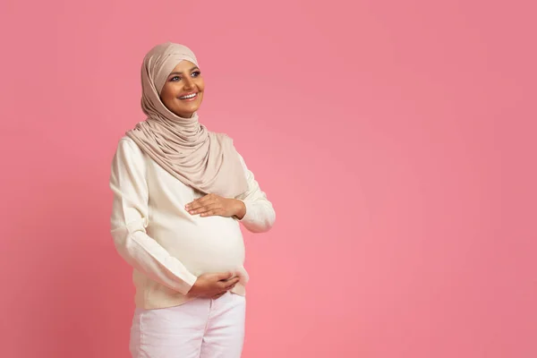怀孕的穆斯林妇女 头戴头巾 抱着肚子 望着别处 等着孩子笑着 爱抚着肚子 期待着中东女人在工作室里摆出粉红的样子 — 图库照片