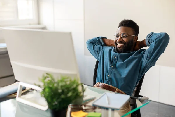 Happy black businessman taking break and enjoying result of project, sitting at table and resting, looking at computer screen, leaning back and holding hands behind head