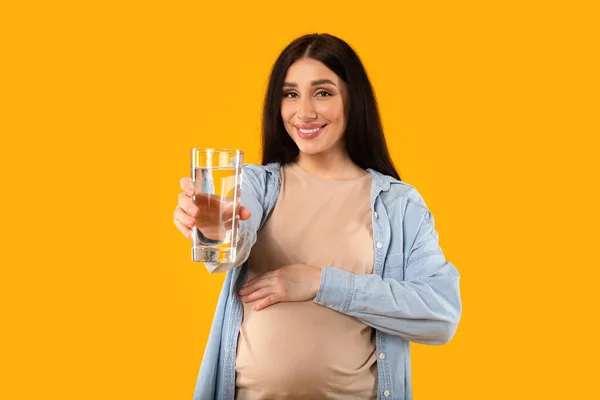 健康的な飲み物だ 妊娠中の女性は腹を受け入れ 新鮮な水でガラスを保持します 美しい期待の女性は さわやかな飲料を提供します 黄色のスタジオの背景に 選択的な焦点 — ストック写真