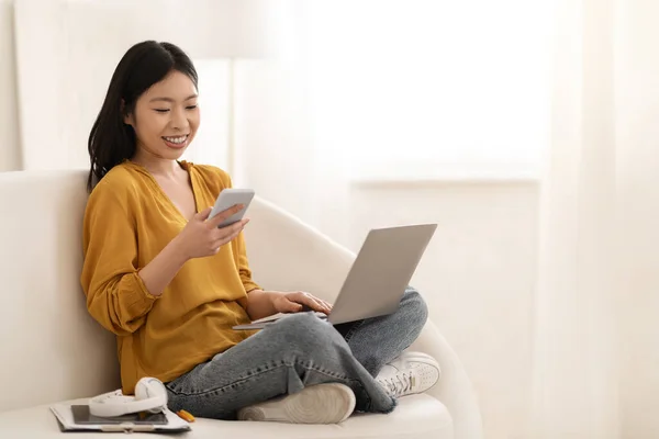 小工具上瘾 积极快乐的日本年轻女性自由职业者 在家工作 女性坐在沙发上 使用笔记本电脑 在智能手机上查看消息 复制空间 — 图库照片