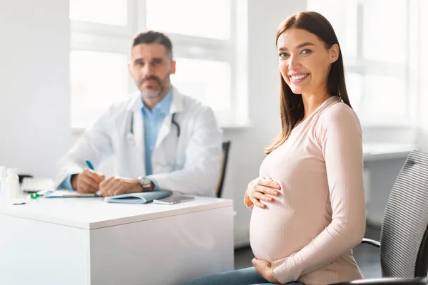 快乐的年轻孕妇在诊所与中年男性妇科医生会诊时坐在一起 抱着肚子 对着镜头微笑 有选择地关注女士 — 图库照片