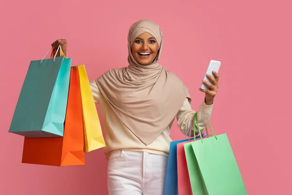 网上购物概念 快乐的穆斯林女性 手持智能手机和明亮的纸袋 伊斯兰女性喜欢季节性销售和网上购物 站在粉色背景上 复制空间 — 图库照片