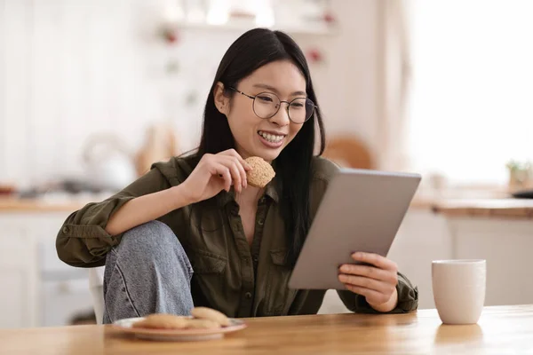 陽気かなり若い中国人女性がカジュアルな服装とアイウェアを着てキッチンテーブルに座って クッキーを食べて デジタルタブレットを使用して ブログを読んで 自宅でコーヒーブレイクを持って スペースをコピー — ストック写真