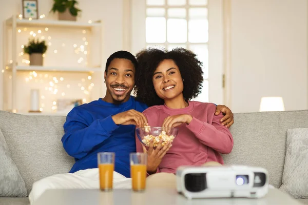 快乐的非洲裔美国人夫妇通过放映机坐在沙发上在家吃爆米花 年轻的配偶拥抱享受电影在线放松的周末 有选择地关注配偶 — 图库照片