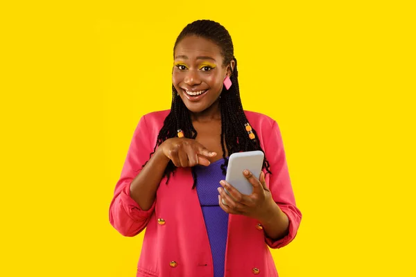 Happy Black Female Pointing At Cellphone Recommending Mobile Application Smiling To Camera Posing Standing In Studio Over Yellow Background. Lady Advertising New App For Smartphone