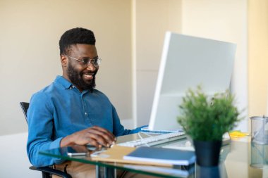 Serbest bir iş. Mutlu siyah adam atölyede bilgisayarla çalışıyor, ofisin içinde oturuyor, boş yer var. Heyecanlı adam cihaz ekranına bakıyor ve gülümsüyor, uzaktan işten zevk alıyor.