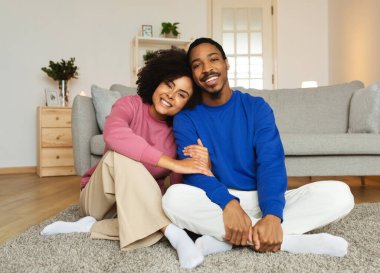 Evdeki modern oturma odasında, yerde oturan kameraya gülümseyen sevimli siyah çift. Genç eşler evde günlük giysiler giyip poz veriyorlar. Mutlu Evlilikler ve İlişkiler