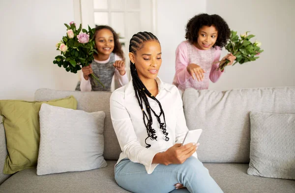 Mothers Day. African American Mommy Texting On Phone While Her Kids Girls Congratulating Her On Holiday, Standing Behind Back With Flowers At Home. Selective Focus On Lady