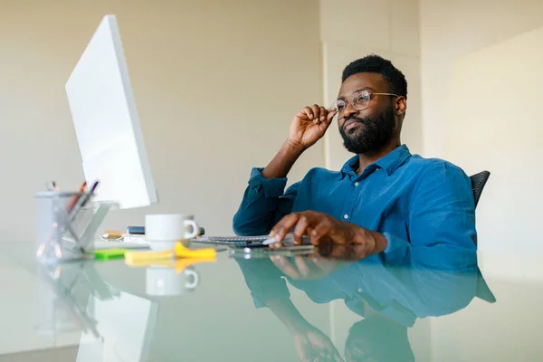 Black businessman sitting at desk in office and working on computer, handsome male entrepreneur reading corporate emails, using computer at workplace, free space