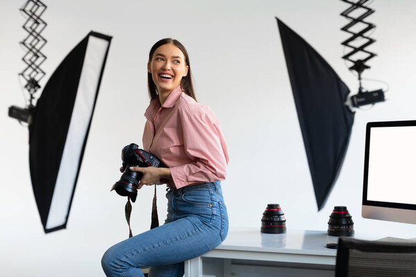 Excited female photographer sitting on edge of table, holding her DSLR camera, looking aside and smiling, working with computer in modern photostudio interior