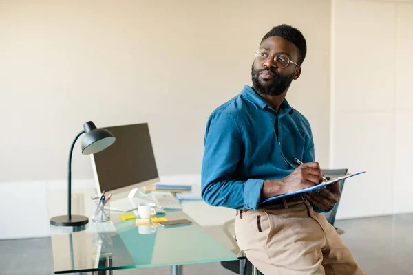 Portrait of thoughtful black man standing in office near workplace, holding clipboard and looking aside at free space, thinking about business project or issues