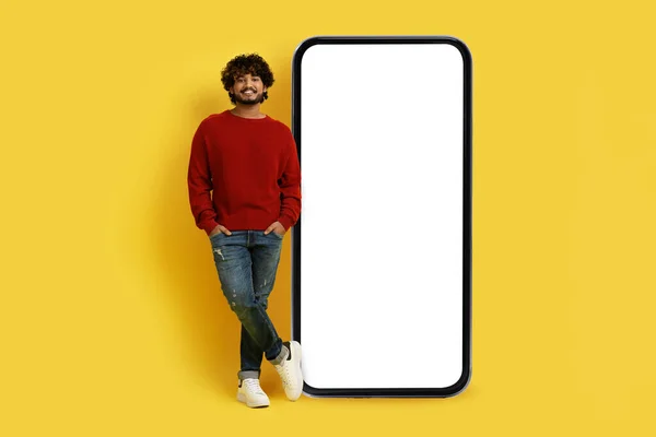 Stock image Positive handsome stylish young indian man standing by huge smartphone with white blank screen, mockup for online offer or mobile app ad, isolated on yellow studio background, copy space