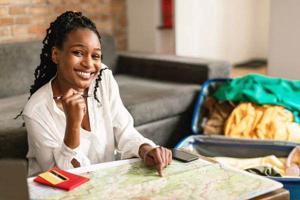 快乐的黑人女人坐在那里 拿着地图和打开的手提箱 计划度假 选择新的国家作为旅游目的地 在客厅对着相机微笑 — 图库照片