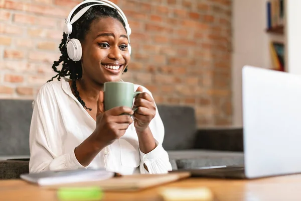 Excited black woman looking at laptop screen and drinking coffee, enjoying remote lecture in headphones, sitting at desk. Female empolyee worker using computer and having break