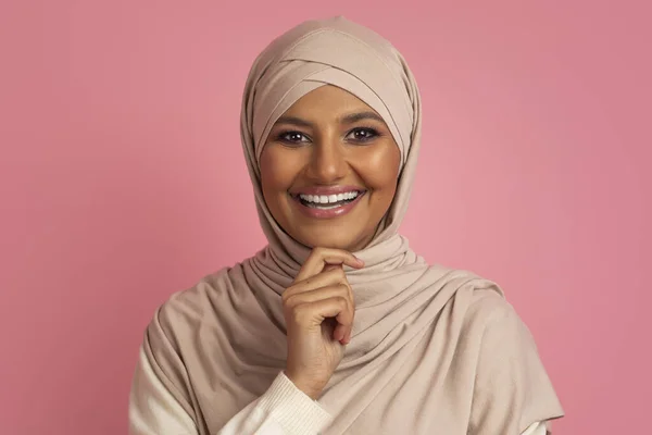漂亮的穆斯林女人 头戴头巾 站在粉红的工作室背景上 在镜头前触摸着下巴 微笑着 快乐快乐的中东女人 伊斯兰之美的概念 — 图库照片