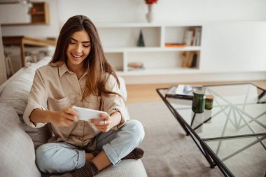 Milenyum melez, ağzı açık, akıllı telefona bakan, oyun oynayan, boş vakit geçiren, oturma odasında tek başına eğlenen bir bayan. Uygulama, evdeki aygıt üzerine video
