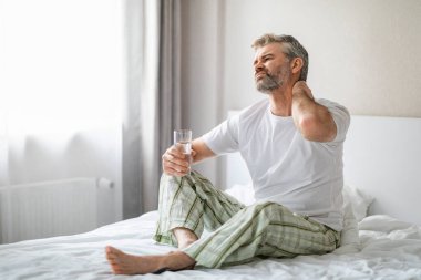 Mutsuz, olgun bir adam, evde yatağında acı içinde, boynunu ovalıyor, sabah kas yorgunluğundan muzdarip, rahatsız bir yatakta uyuduktan sonra, elinde bir bardak su tutarak, fotokopi odasına bakıyor.