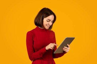 Turuncu stüdyo arka planında modern dijital tablet kullanan kırmızı dijital göçebe bir genç kadın. Ped ekranına dokunuyor ve gülümsüyor. Güzel uygulamalardan keyif alıyor.