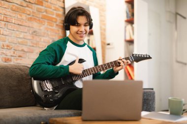 Asyalı ergen çocuk dizüstü bilgisayarda gitar çalıyor, parmaklarını hareket ettiriyor, koltukta oturmayı öğreniyor, kulaklık takıyor. Müzikal hobi ve eğlence. Seçici Odaklanma