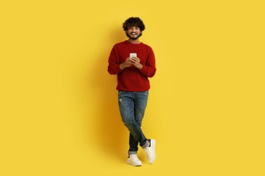 Yakışıklı, kıvırcık, bin yıllık Hintli adam sarı stüdyo arka planında cep telefonuyla poz veriyor, kameraya gülümsüyor, güzel mobil uygulama kullanıyor, tam boy, kopyalama alanı kullanıyor.