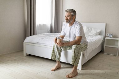 Mutlu, neşeli, dinlenmiş, çekici, gri saçlı, pijama giyen, evde çıplak ayaklı, elinde fincanla oturan, çay ya da kahve içen, fotokopi odasına bakan olgun bir adam.