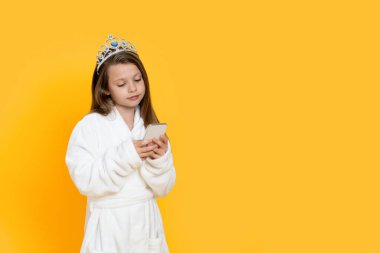 Üzerinde bornoz ve oyuncak taçla gezen, interneti kolaçan eden ya da internetten alışveriş yapan, sarı arka planda tek başına duran, uzay kopyalayan sevimli küçük kız.