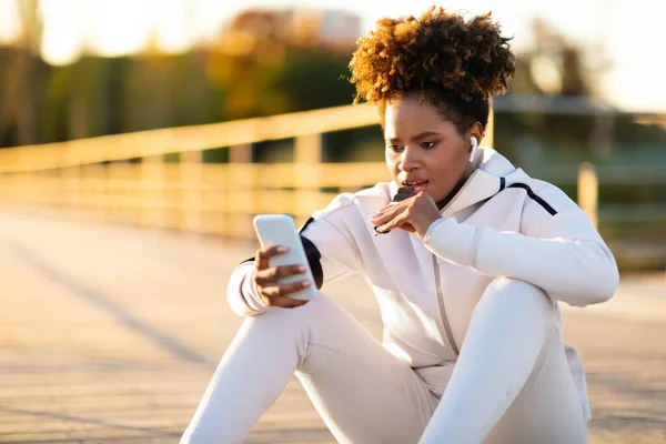 Young Sporty Black Woman Eating Protein Bar Using Smartphone Outdoors — Stock Photo, Image