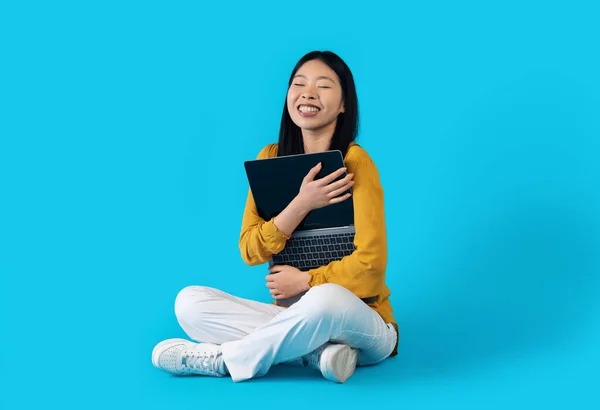 Gadget addiction. Overjoyed happy beautiful young korean woman hugging open laptop with blank screen and smiling, sitting on floor over blue studio background, copy space