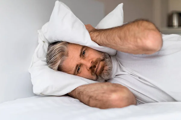 Portrait of unhappy exhausted middle aged man lying in bed and covering ears with pillow, hearing and suffering from too loud sound or snoring, tired of noisy neighbors. copy space