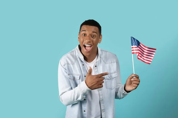 Cheerful shocked excited millennial black guy in casual pointing finger at USA flag, recommend study, isolated on blue studio background. Learning english, exchange education, great offer