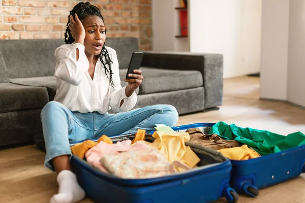 旅行の問題の概念 携帯電話を使用してショックを受けた黒人女性と衝撃的な旅行のニュースを読んで 自宅で満員のスーツケースの近くに座っている 飛行機はキャンセルだ — ストック写真