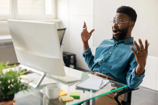 Got job promotion. Excited black businessman celebrating great news and luck, looking at computer screen with open mouth, sitting at workplace in office interior