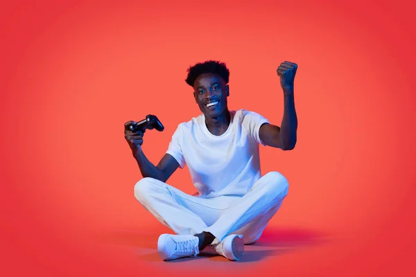 喜びに満ちた若いアフリカ系アメリカ人男性で 白いカジュアルな服装で床の上に座っているネオンライトの赤いスタジオの背景に ビデオゲームをプレイし ジョイスティックを保持し 勝利を祝う スペースをコピー — ストック写真