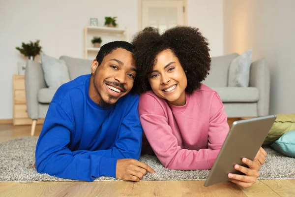 Cheerful Black Couple Using Digital Tablet Browsing Internet And Smiling To Camera Lying On Floor At Home. Spouses Posing With Computer Websurfing Together On Weekend. Technology And Gadgets