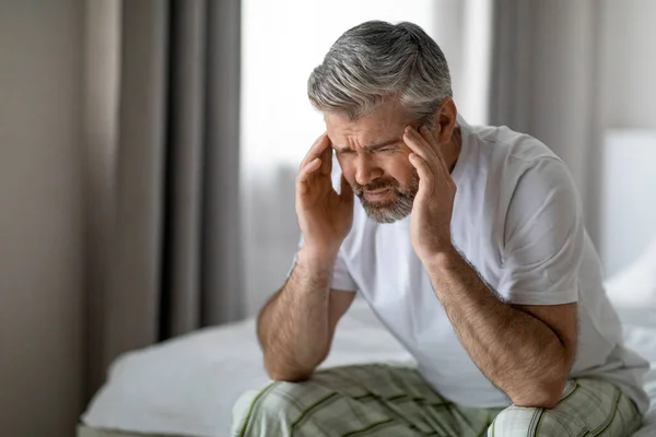 Closeup of unhappy middle aged handsome grey-haired bearded man in pain sitting on bed, touching his head, suffering from headache, migraine, hangover, white bedroom interior, copy space