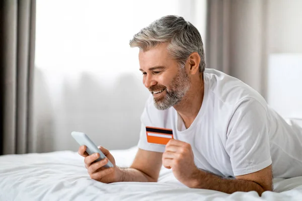 E-commerce, retail. Cheerful handsome middle aged man with beard lying on bed, using smartphone and plastic bank card, making online order while staying home at weekend, copy space