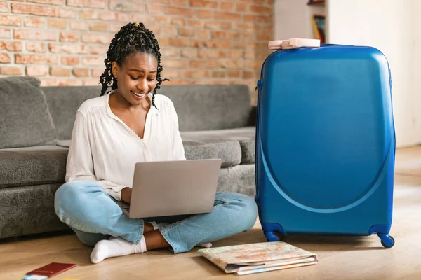 快乐的黑人女人计划度假旅行 在网上使用笔记本电脑预订酒店房间 坐在家里的行李箱旁 兴奋的女性游客在电脑上浏览出租度假房地网站 — 图库照片