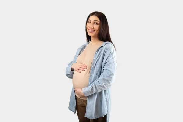 可愛い妊婦待っている赤ちゃん カジュアルな服装でポーズ カメラを見て笑顔 白いスタジオの背景に隔離されたおなかを受け入れます フリースペース — ストック写真